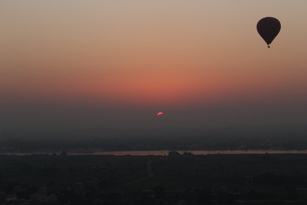 Sun rising in the horizon and, in the foreground, a hot air balloon flies over the Nile, in Luxor