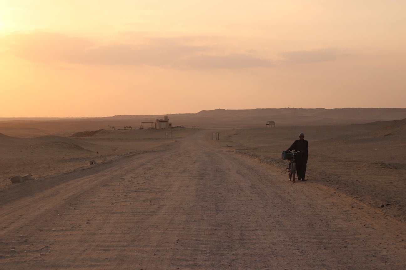 Sunset in the Saqqara complex, a man walks with his bike through the long path to the exit